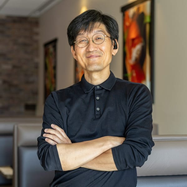Smiling Asian man in glasses and a black polo shirt with arms crossed, standing in Sushi Hana.
