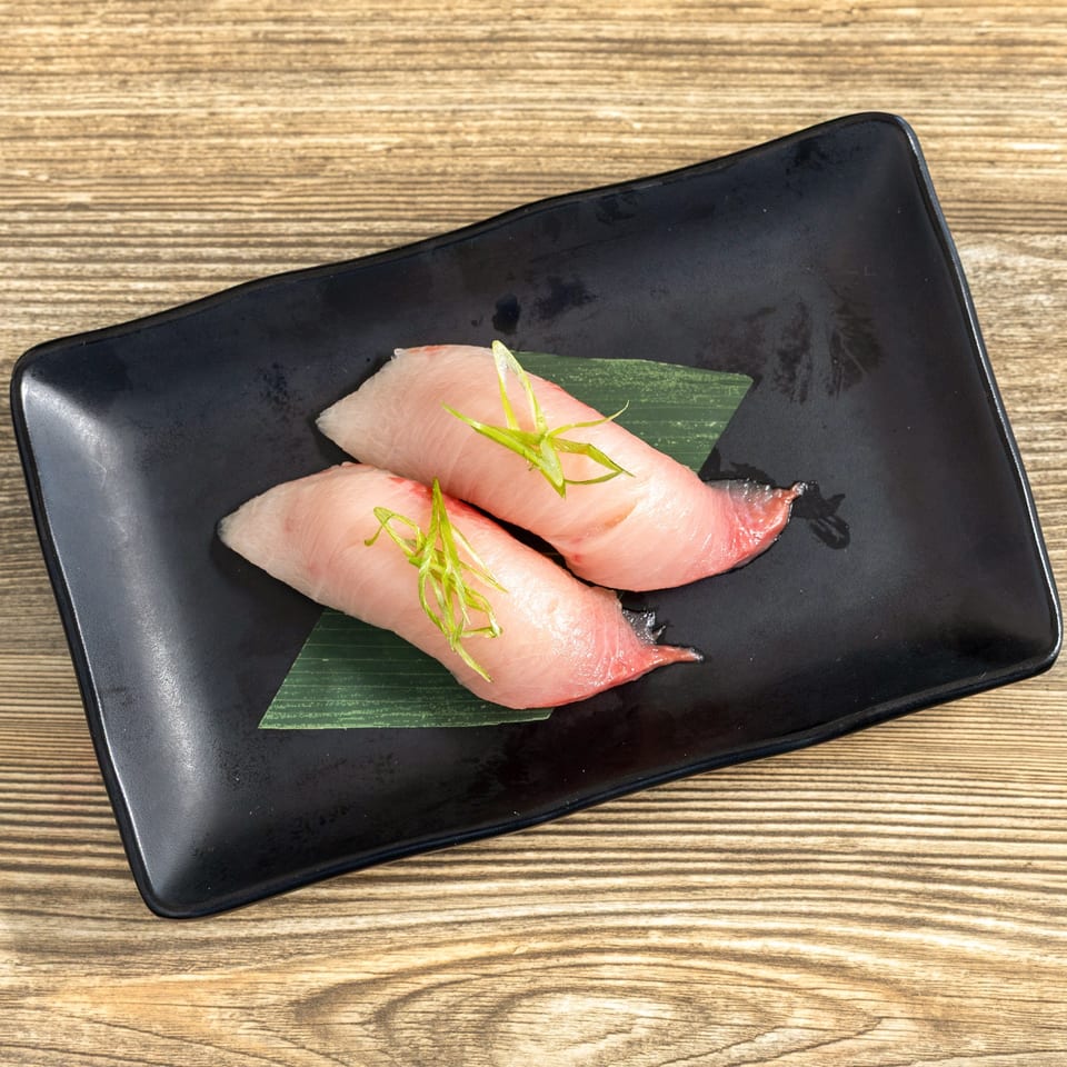 Two pieces of Hamachi sushi, topped with green onions, on a black plate with a bamboo leaf.