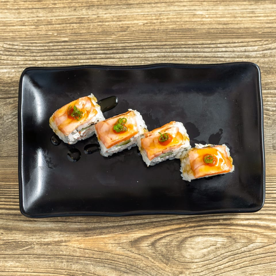 Four pieces of Sushi Hana salmon sushi rolls with sauce and green garnish on a black plate.