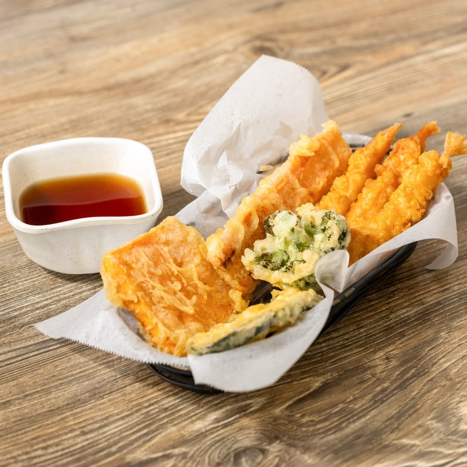 Crispy tempura shrimp, fish, and vegetables in a basket with a side of dipping sauce.