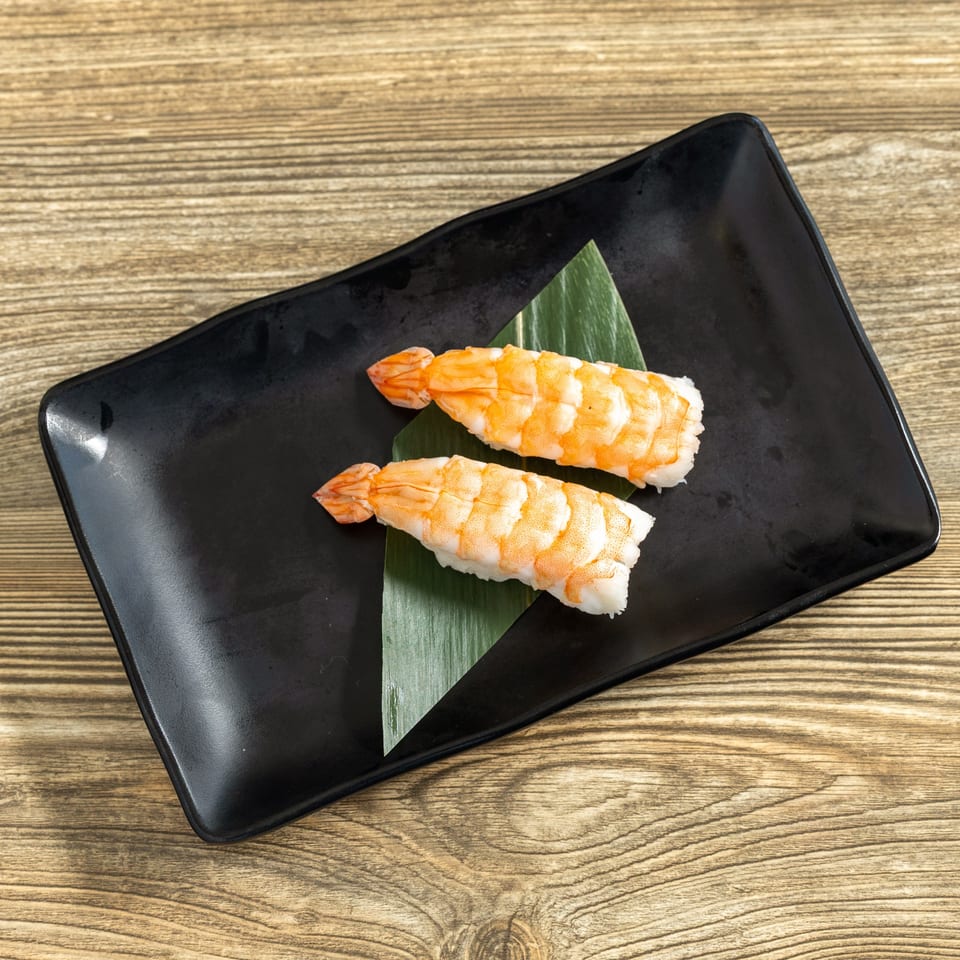 Two pieces of shrimp nigiri sushi on a black rectangular plate with a green leaf.