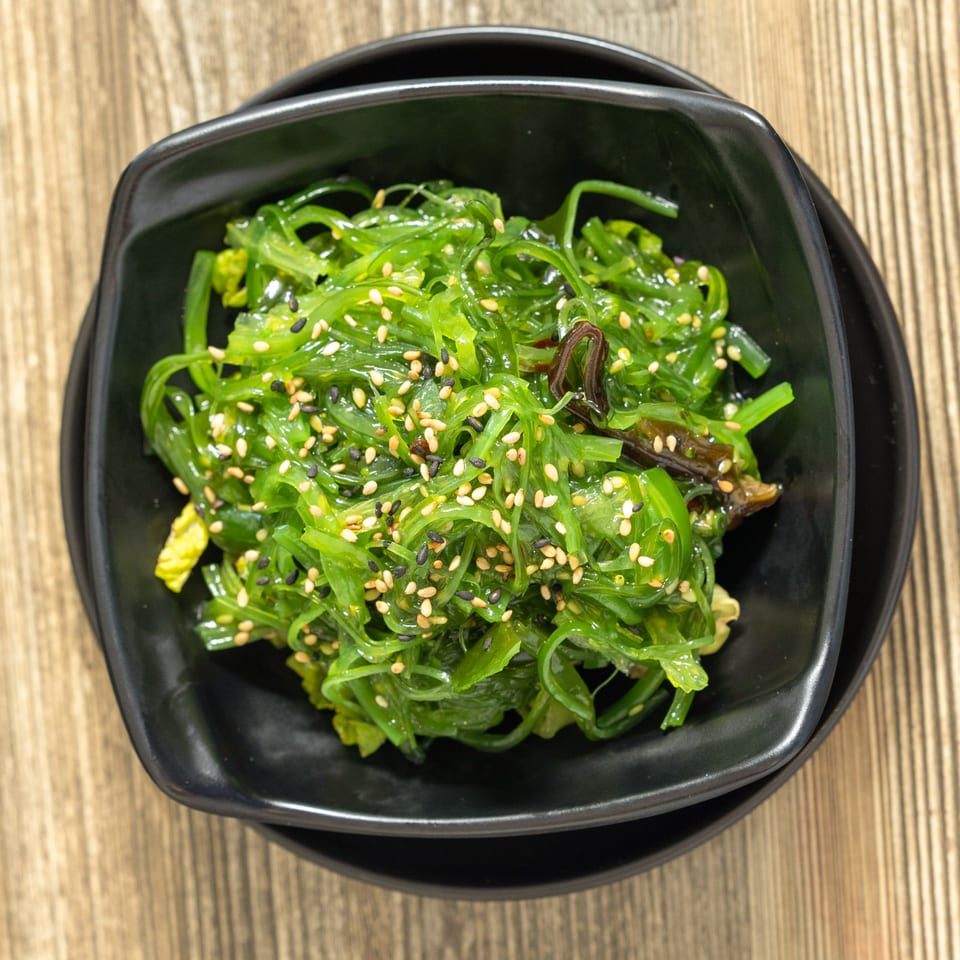 Sushi Hana's vibrant green seaweed salad, topped with white and black sesame seeds, in a black bowl.