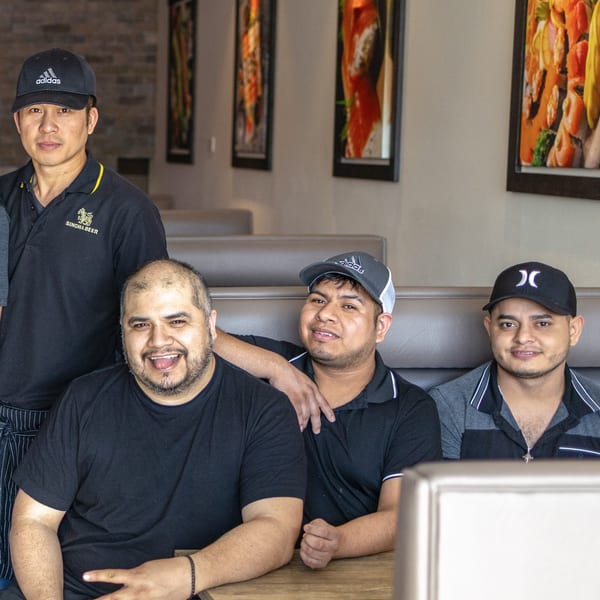 Four male Sushi Hana staff members, three seated in a booth, one standing, smiling at the camera.